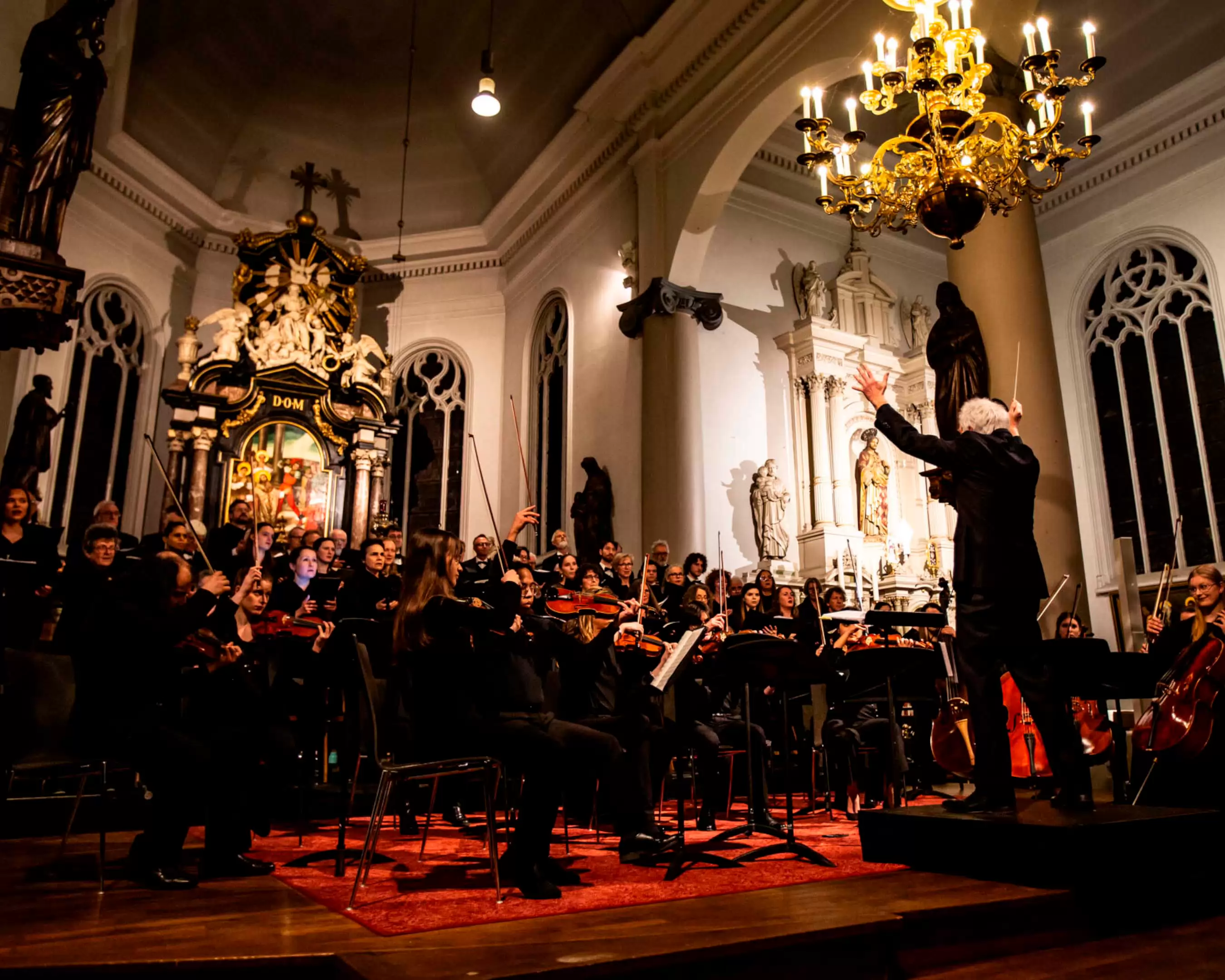 An orchestra performs a musical piece in a church. In front of the group is a conductor. String instruments and singers can be seen. 