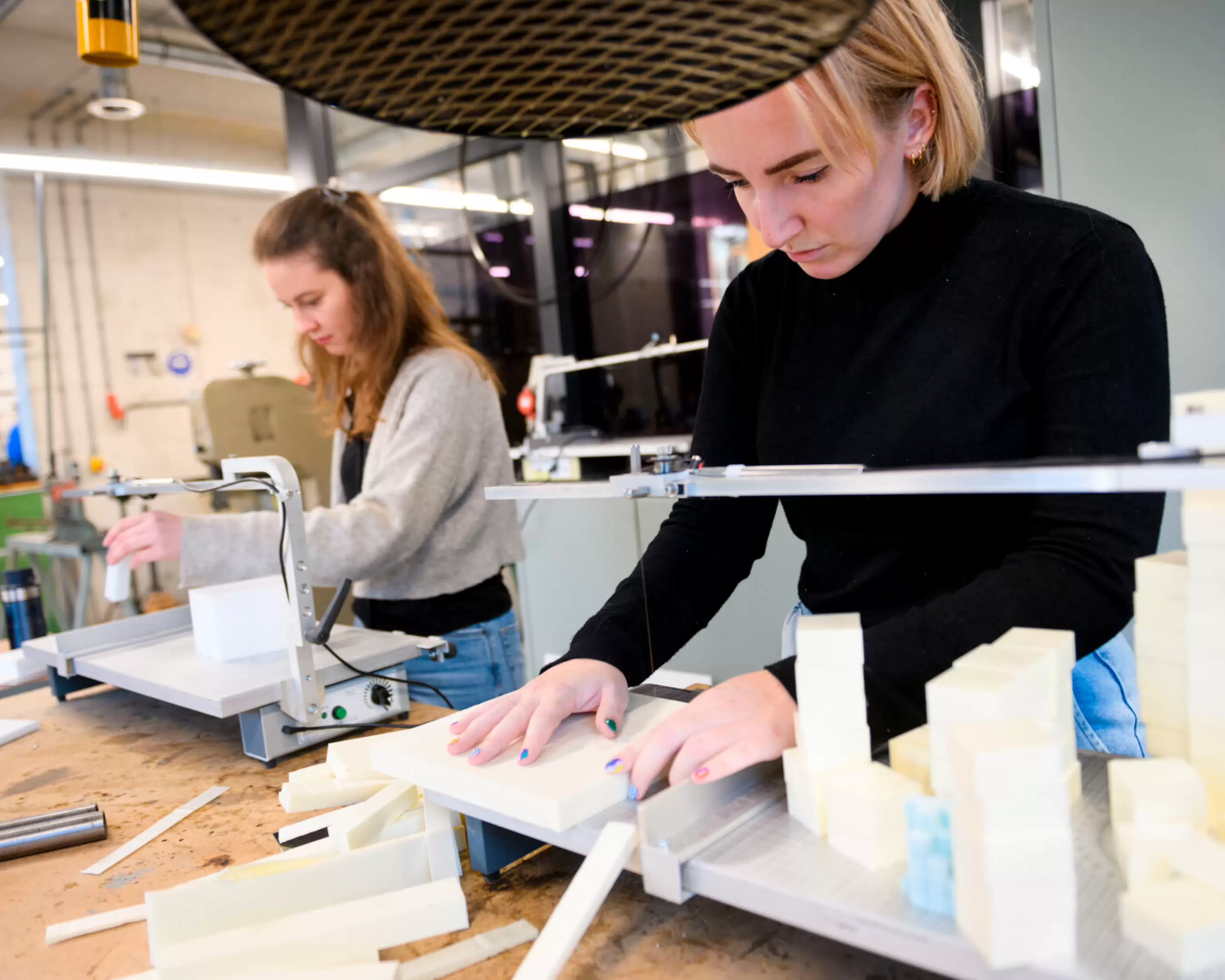 Two students from the Master's program in Urbanism working in the wood workshop. They are using precision cutting tools to shape foam blocks, likely for architectural models. The workspace is equipped with various tools and materials, creating a hands-on learning environment.