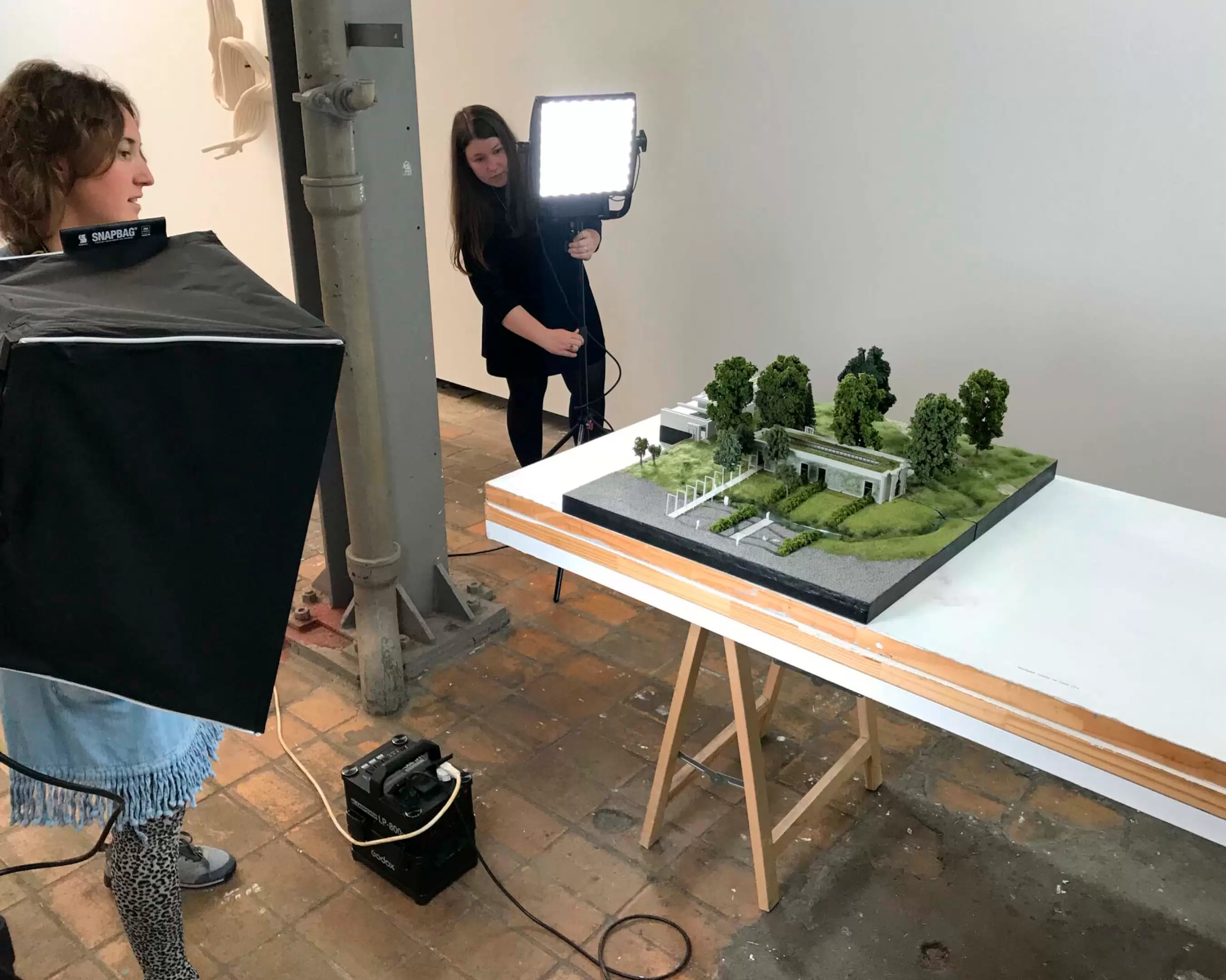 Two students from the master Architecture are photographing there scale model project. The scale model consists of green grass and trees with a grey building 