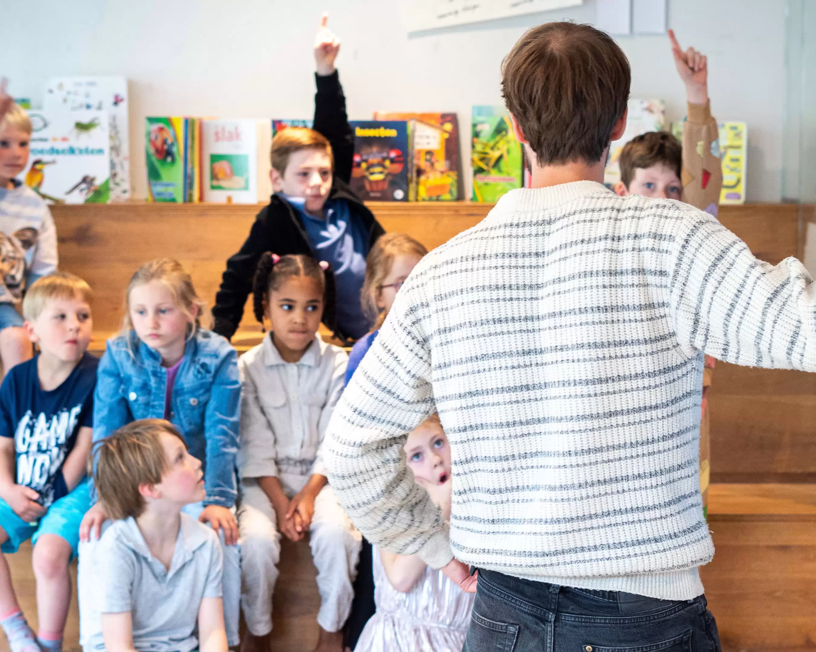Docent voor de klas, waarbij de leerlingen in een theaterachtige opstellen luisteren en hun vinger opsteken.