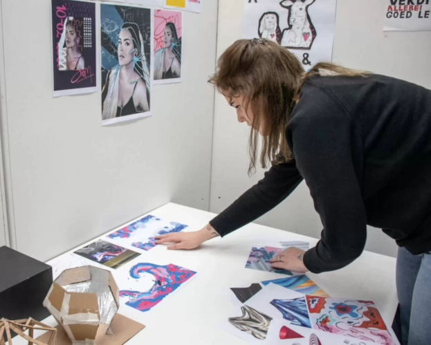 Student bekijkt eigen kunstwerken op tafel, met posters aan de muur.