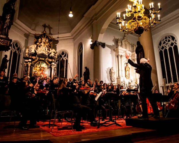 An orchestra performs a musical piece in a church. In front of the group is a conductor. String instruments and singers can be seen. 